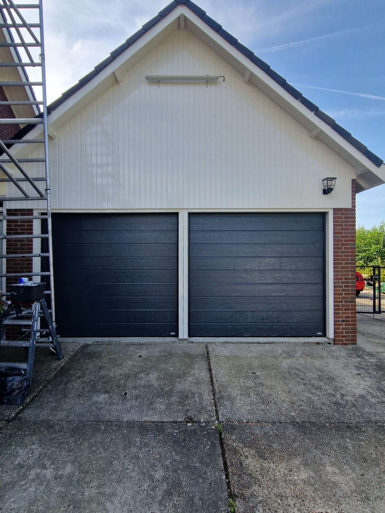 Wit geschilderde houten topgevel boven twee antraciet garagedeuren, met een rolsteiger ernaast