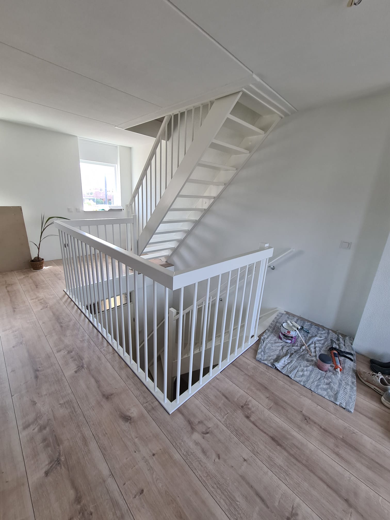 Wit geschilderde open trap met balustrade in een lichte kamer, met afdekkleed en verfblik op de vloer